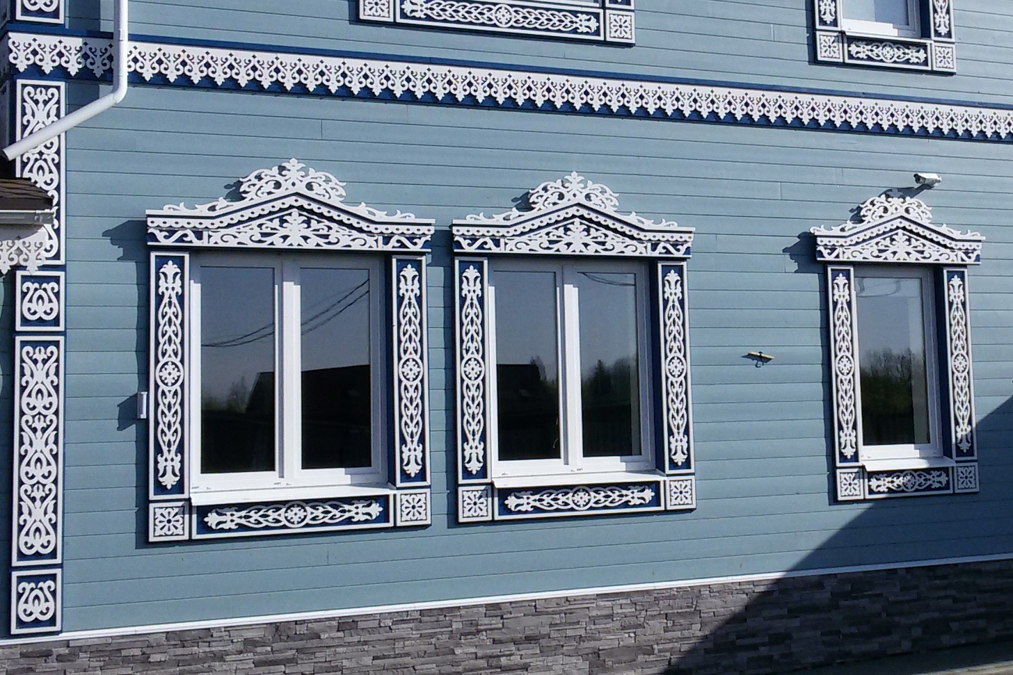 Монтаж светлых резных наличников и карнизов на голубом деревянном фасаде